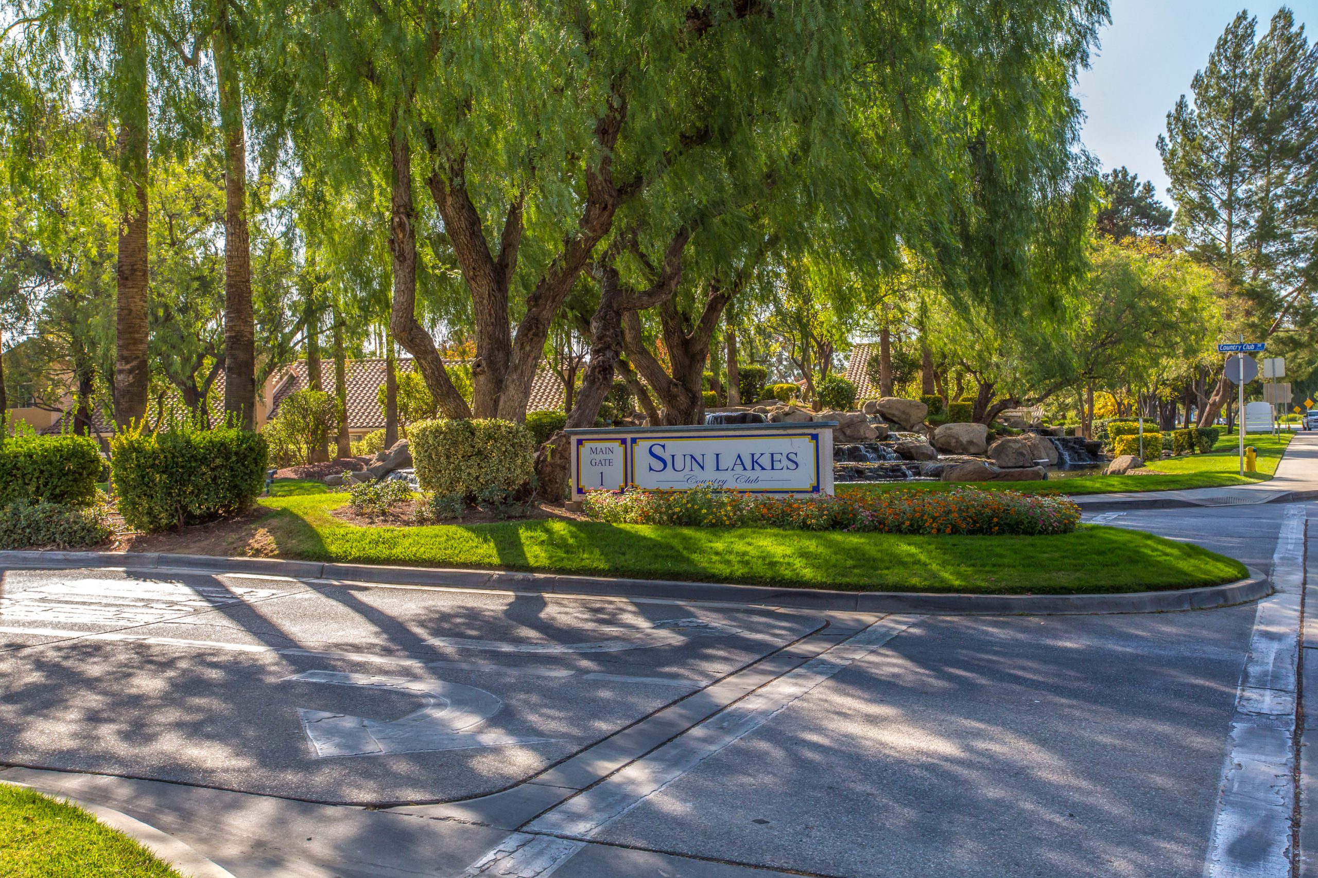 Entrance to Sun Lakes Country Club in Banning, CA, featuring landscaped greenery, a gated entry, and directional signage for visitors.